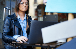 woman in a black jacket sitting at a table and typing on a laptop