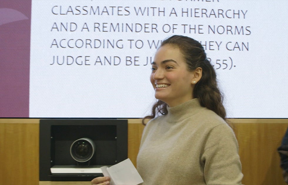 Student smiling standing in front of a screen that reads &quot;...classmates with a hierarchy and a reminder of the norms according to...can judge and be...&quot;