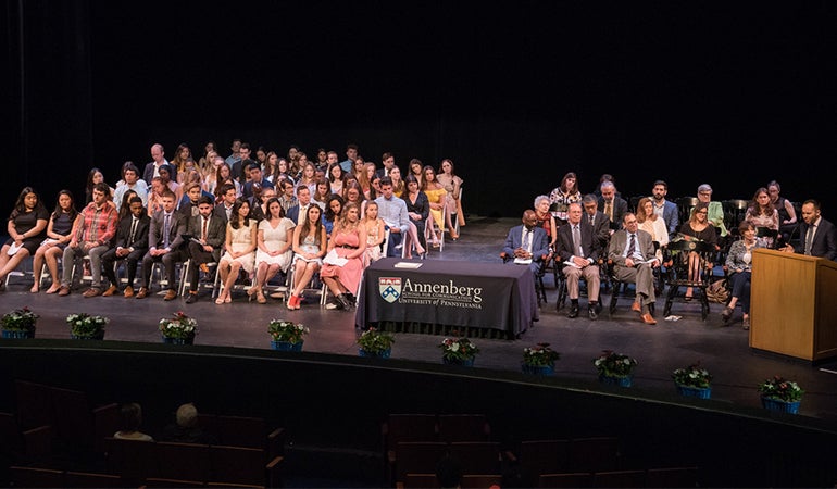 Graduates and faculty seated in chairs on a stage