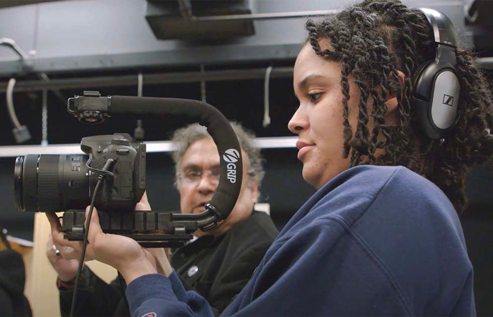 Student looking into the lens of a camera rig while an instructor looks on