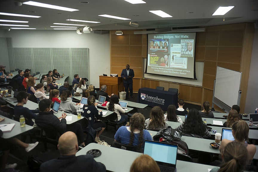 Eric Deggans lecturing students with a powerpoint. The current slide has the title "Building Bridges not Walls", under which is a screenshot of a news channel with 4 people talking via video, a screenshot of a site with an article related to Bill Clinton, a screenshot of a Donald Trump tweet, and a Daily News Magazine cover with Donald Trump's face with headline "Had eyes for a 10-year-old". Below is "Decoding Media's Confusing Coverage of Race, Gender & Culture This Election Season". 