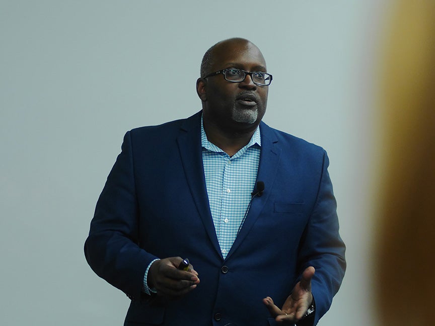 Eric Deggans photographed candidly while taking. He is wearing a navy blue suit jacket and a light blue and white striped shirt.