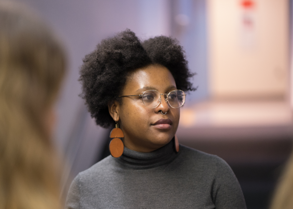 Florence Madenga looking to her left. She wears glasses, long brown earrings and a grey turtleneck.