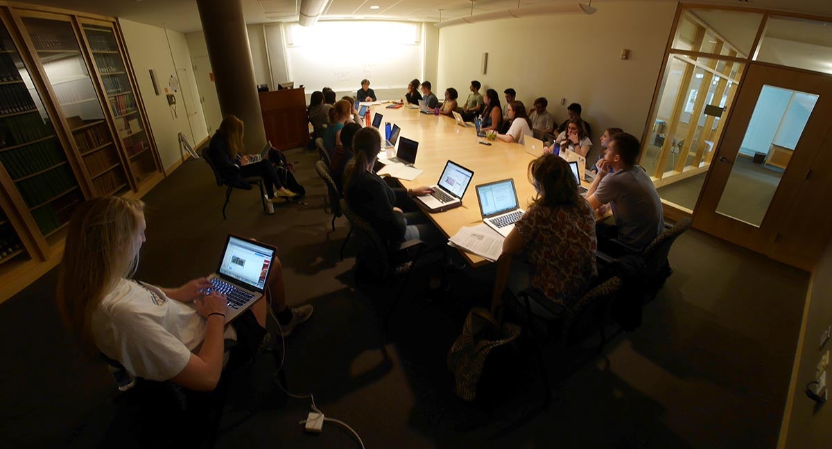 Dim classroom with a table filled with students on laptops as they listen to Joan Garry who is at the front.