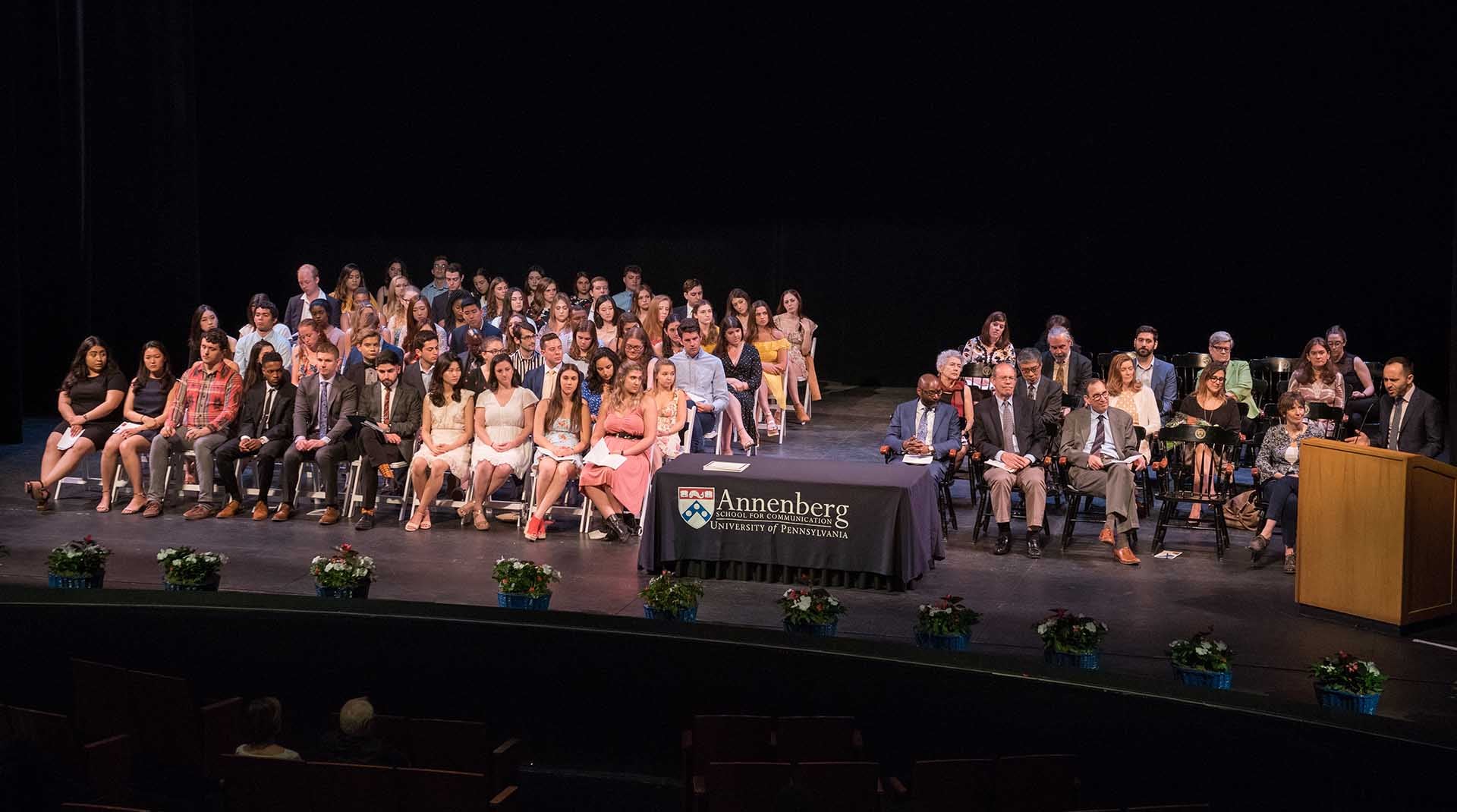 aerial view of the stage during graduation. A large number of students are seated in folding chairs, and faculty are on the right in folding chairs. A speaker is at the podium.