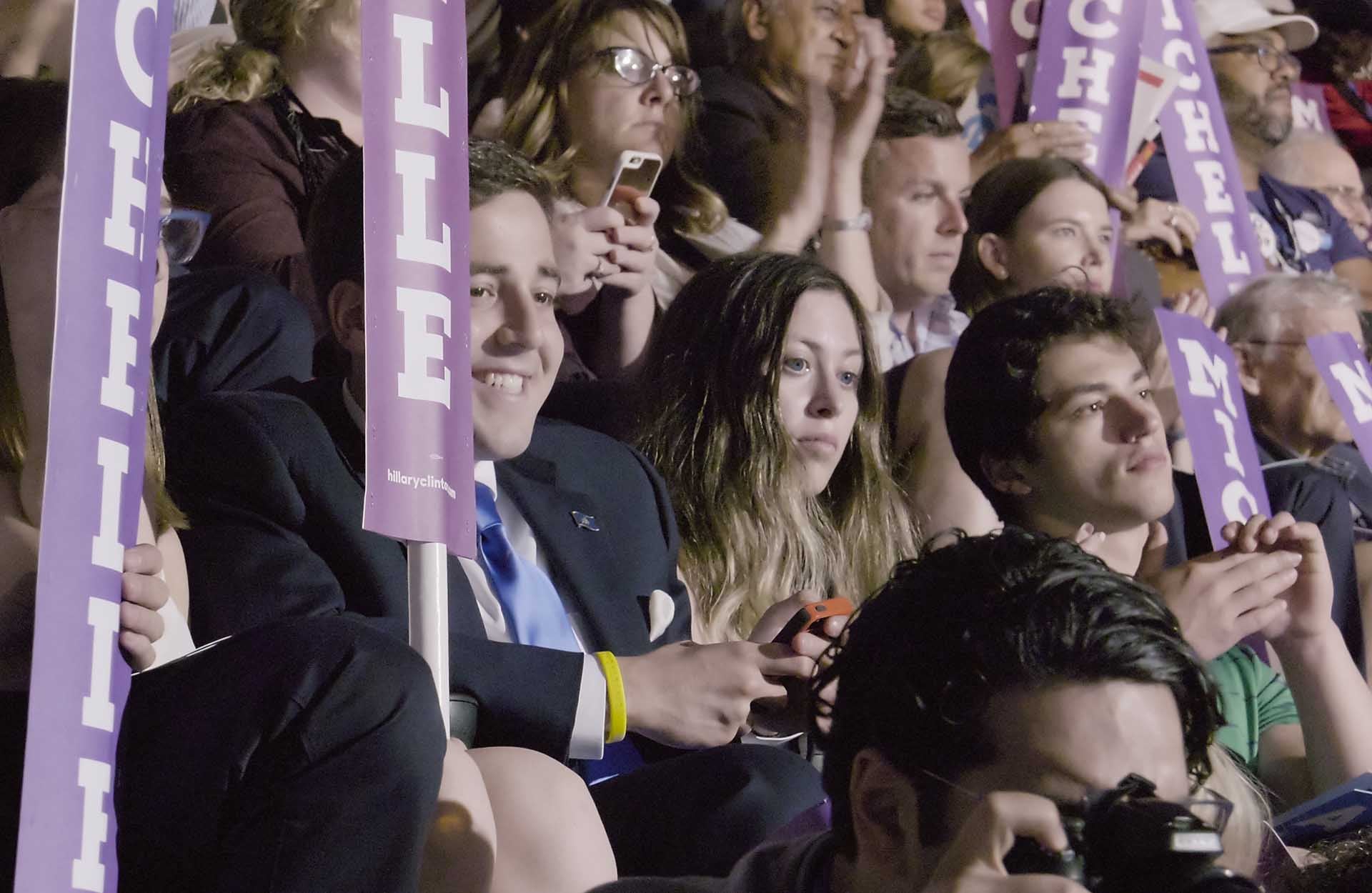 still01 1920x1250 CROP.jpg A crowd of people looking in front of them. Some are holding up long, purple signs with white text that says 'Michelle' in upper-case letters.