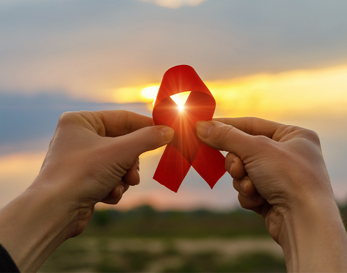 ApplicationstoHealth_Checkerboard.png Hands holding up a red HIV ribbon in front of a sunset