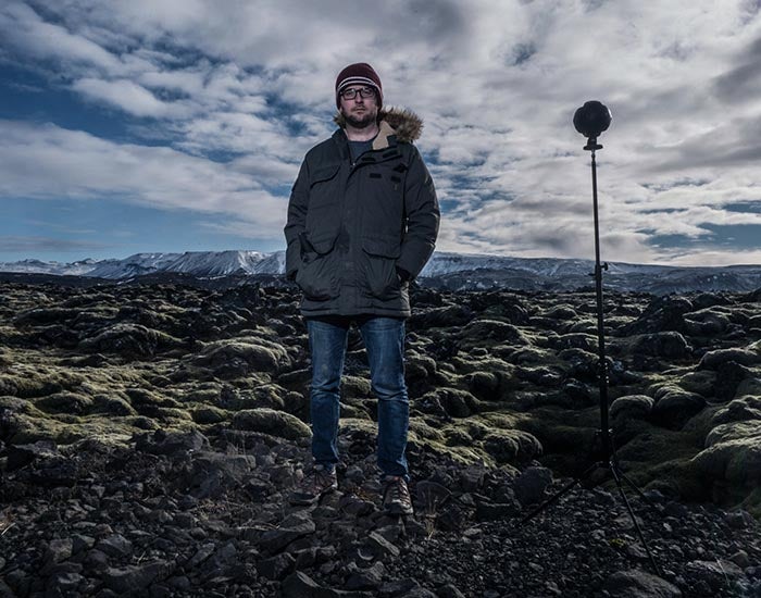 Person in winter gear stands outdoors in an eerie-feeling landscape with rocks and plants and dramatic clouds in the sky. A VR camera on a tripod also stands in the foreground