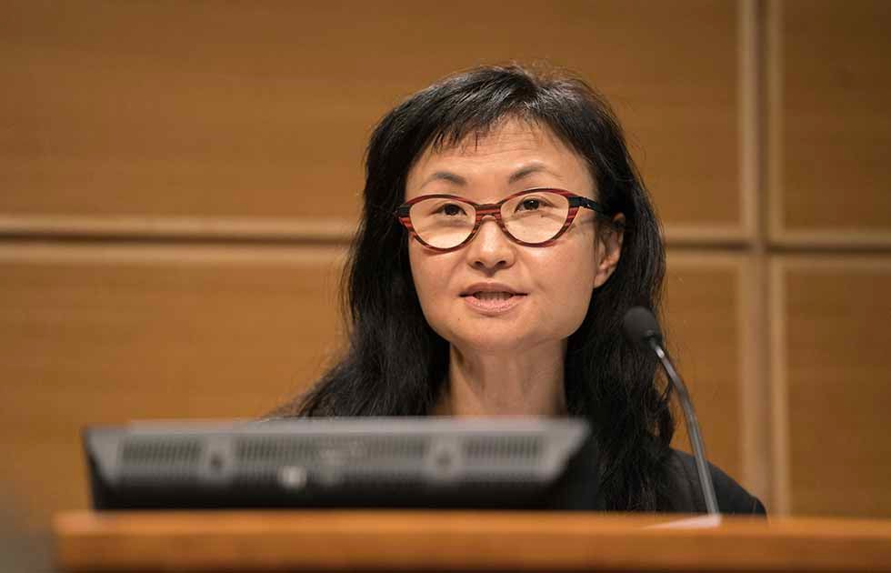 Wendy Chun standing behind a podium speaking with a wood-paneled wall behind her