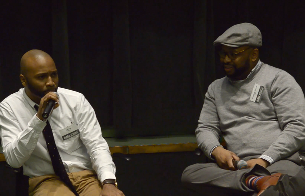 Two people sitting in chairs in front of a black background. One is speaking into a microphone while the other looks at the speaker.