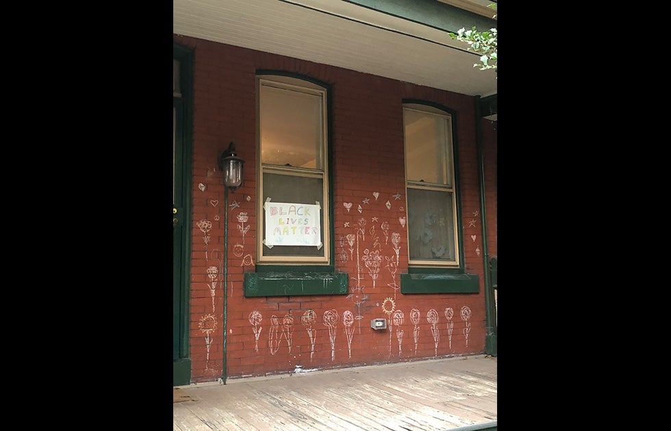 Brick building with a colorful hand-written sign (appears to be made by a child) that says “Black Lives Matter” and chalk art on the brick facade that is mostly flowers, hearts, peace signs, and stars.