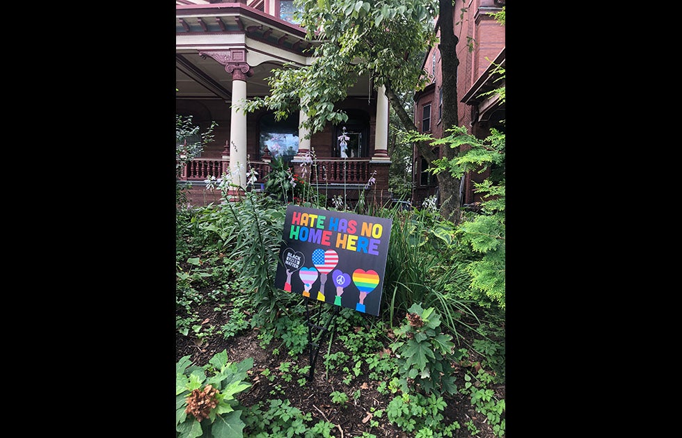 Yard sign that says “Hate has no home here” with multiple hands (of different skin tones) holding hearts. One heart says “Black Lives Matter” one is a trans flag, one is an American flag, one has a piece sign, and one is a rainbow. The letters are in a rainbow pattern. It does not appear to be handmade.