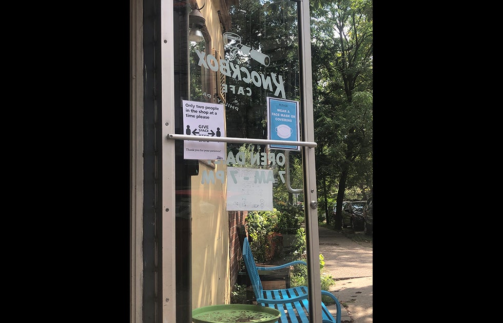 Window of Knockbox Cafe that has two signs. One says “Only two people in the shop at a time please” with cartoons of people distanced and “Please wear a face mask or covering” with an image of a mask.