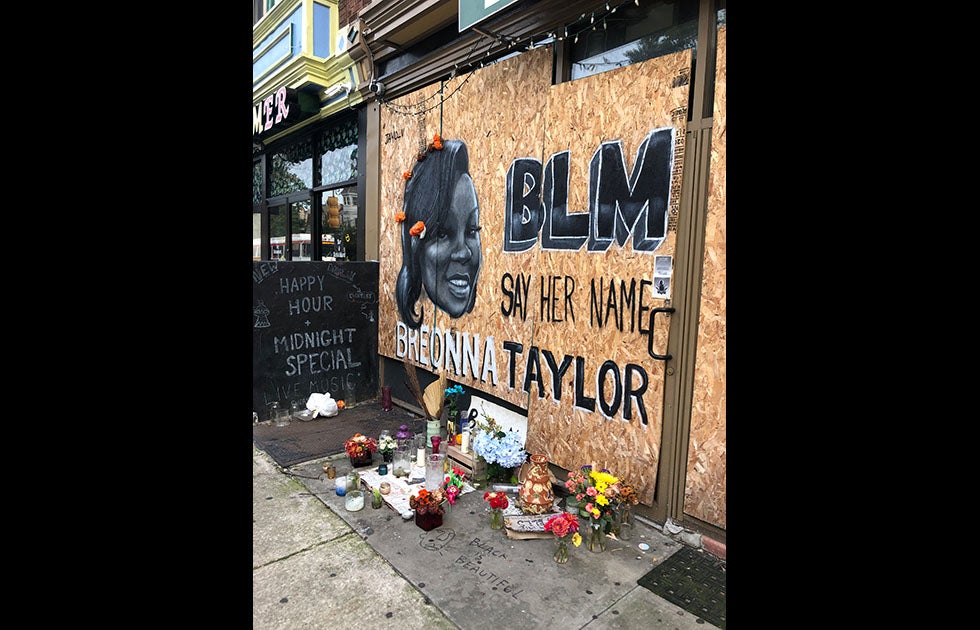 A memorial for Breonna Taylor. It is painted on a boarded up building and reads “BLM Say Her Name Breonna Taylor” in black and white and has a portrait of her. Her hair has real flowers attached to it. There are flowers and candles below the memorial forming an altar. The sidewalk has Black is Beautiful painted on it with an abstract face next to it (same as featured on the dumpster).