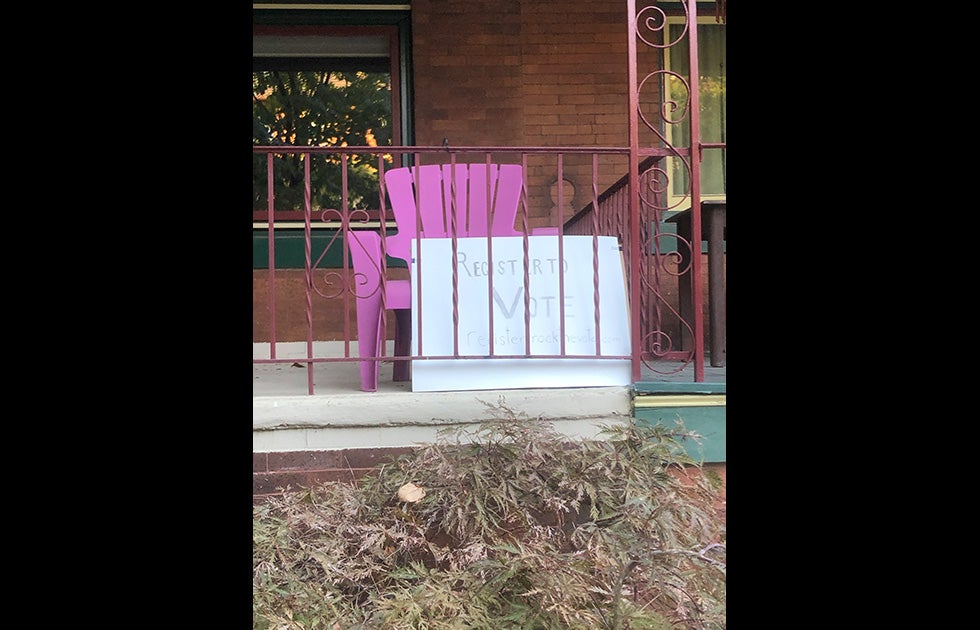 Handwritten poster board on a porch that says “Register to Vote” and has a URL partially obstructed by the balcony fence.