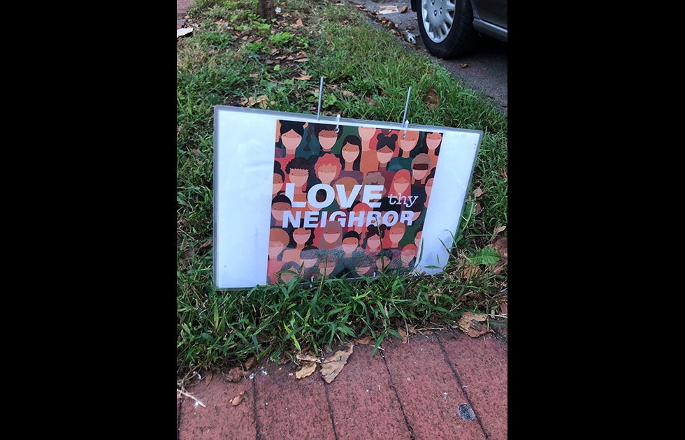 Laminated sign that says Love thy neighbor and has cartoon pictures of people of various skin tones wearing masks around their noses and mouths.