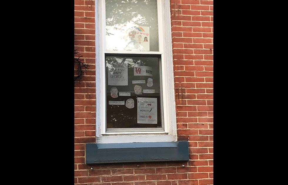 An apartment window with multiple small, handmade signs. One says “Breonna, Tony, George” and one says “LOVE not Hate” and one has a rainbow and hearts with drawings of people. The rest are not legible.