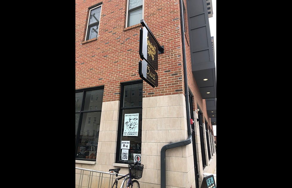 Crust Vegan Bakery bakery with multiple signs in window. Two are not legible. One says “Hours 9-4” and another reads “Defund Police/All Resources to Community” with an image of a hammer breaking a piggy bank wearing a police uniform.