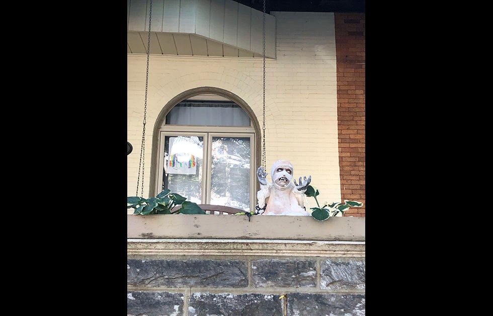 Balcony with porch swing and a Halloween decoration of a mummy. There is also a sign in the window that appears to be made by a child and is very colorful.