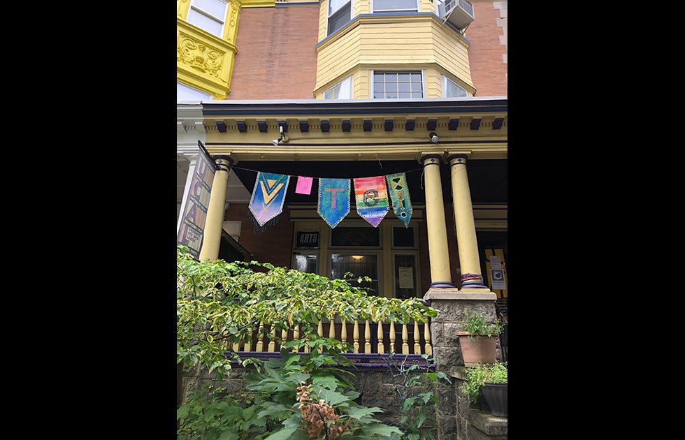 Flags hung outside of an arts and dance studio that say “VOTE” on various pieces of fabric in different shapes and colors. The “o” is a smiley face and the. E is on a rainbow flag.