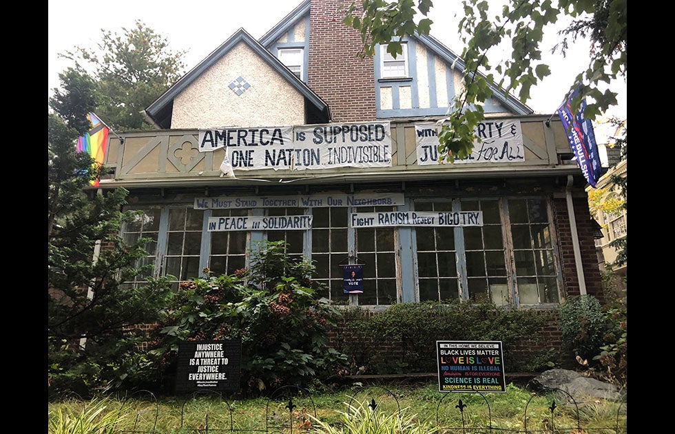 House with many signs and flags; some handmade, some not. One flag says “No more Bullshit” under something else that is not legible in the picture. Another has the Philadelphia LGBTQ pride flag. One of the large handmade banners reads “America is supposed” then “One nation indivisible” with one piece folded down (presumably saying “be”).