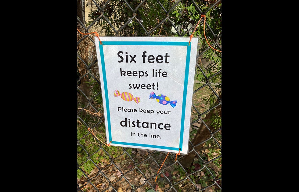 Chain link fence with a sign that says “six feet keeps life sweet! Please keep your distance in the line” with two pictures of candy.