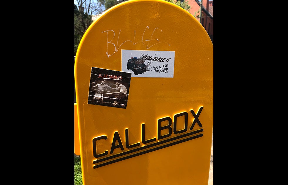 A yellow call box with two stickers on it. One is tattered and shows boxers with the text “Blak Madeen” and another is more pristine and says “2020 blaze it still not loving the police” with an image of a cartoon police car on fire.