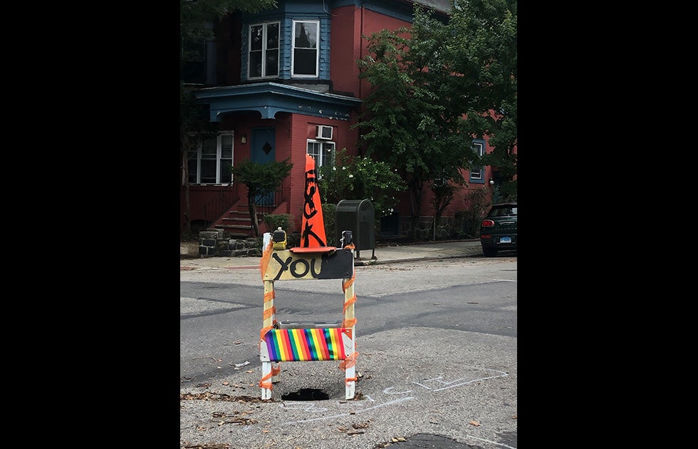 a small sink hole with a traffic barrier and a pylon on top. It is spray painted with “FUCK YOU” and there is rainbow tape wrapped around it.