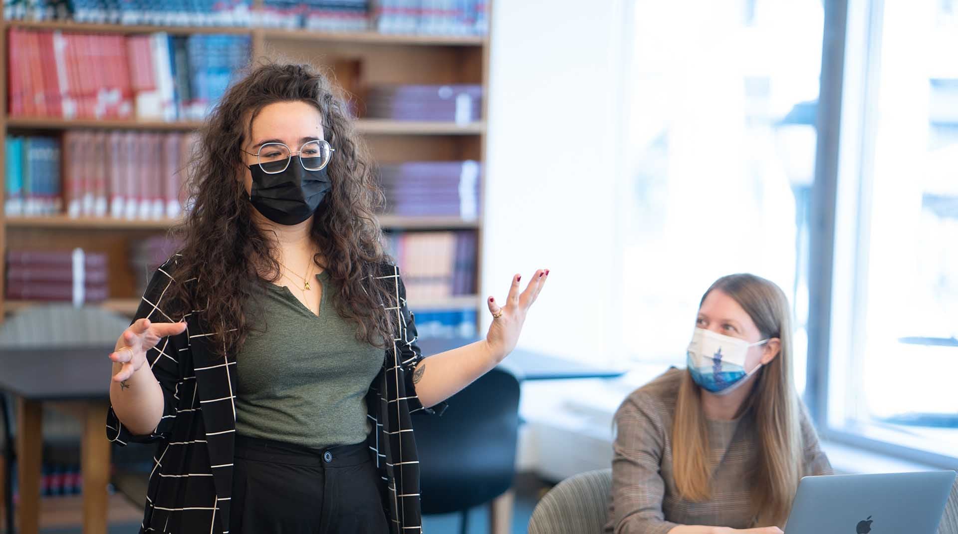 Woman gesturing and speaking while she stands in the library. Another woman looks on.