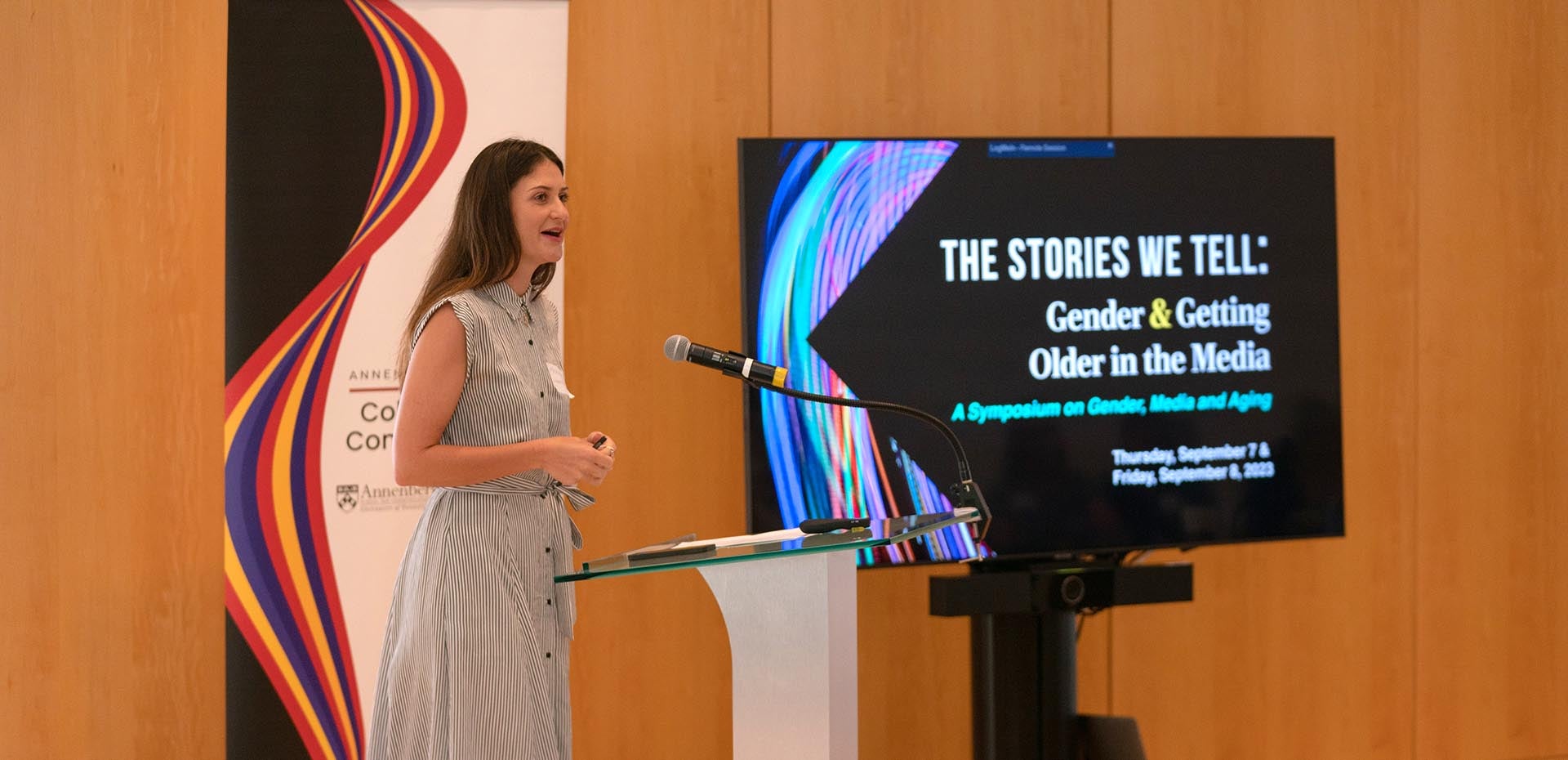 Woman standing at a podium with a banner behind her and a TV screen with the words "The Stories We Tell: Gender & Getting Older in the Media"