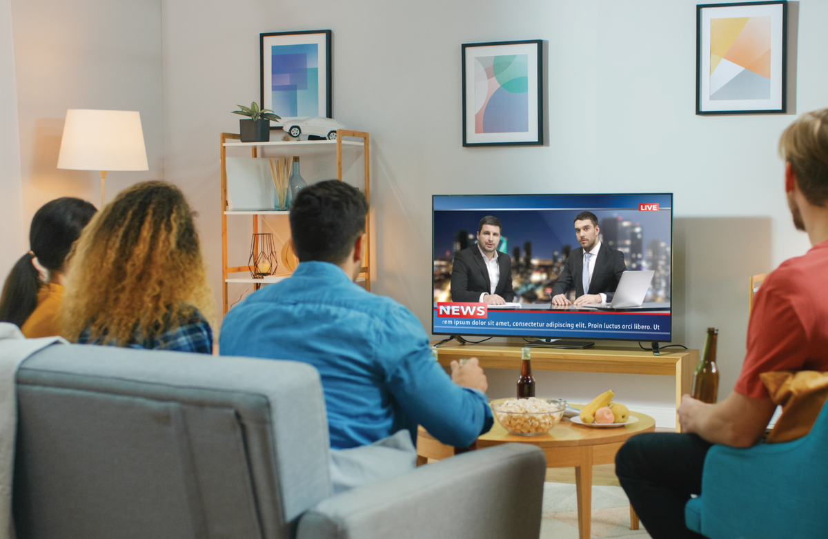 Group of young adults watching a live news broadcast in a modern living room, with snacks and drinks on the coffee table.