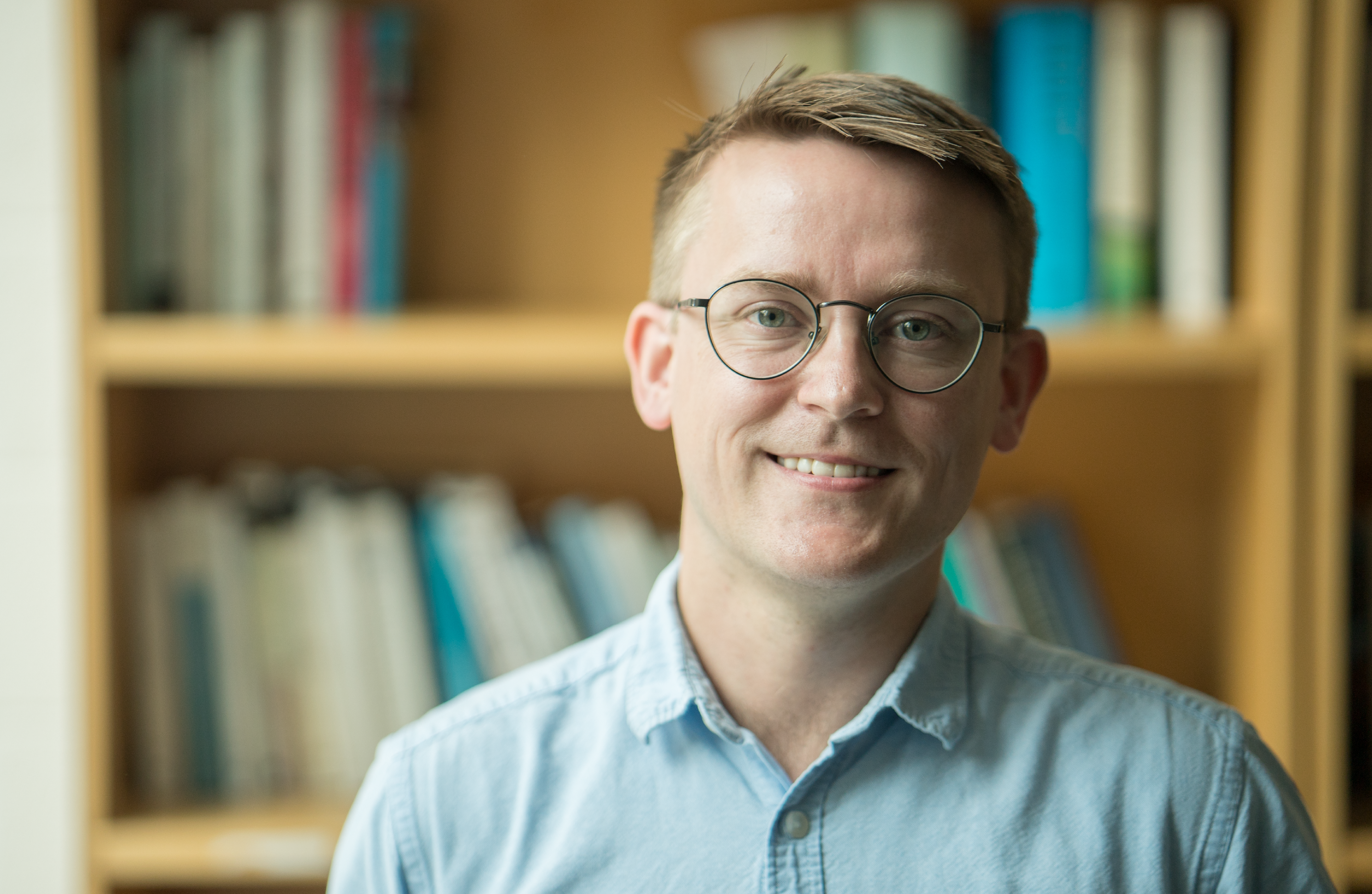 t: David Lydon-Staley, wearing round glasses and a light blue button-up shirt, smiling in front of a wooden bookshelf filled with books.