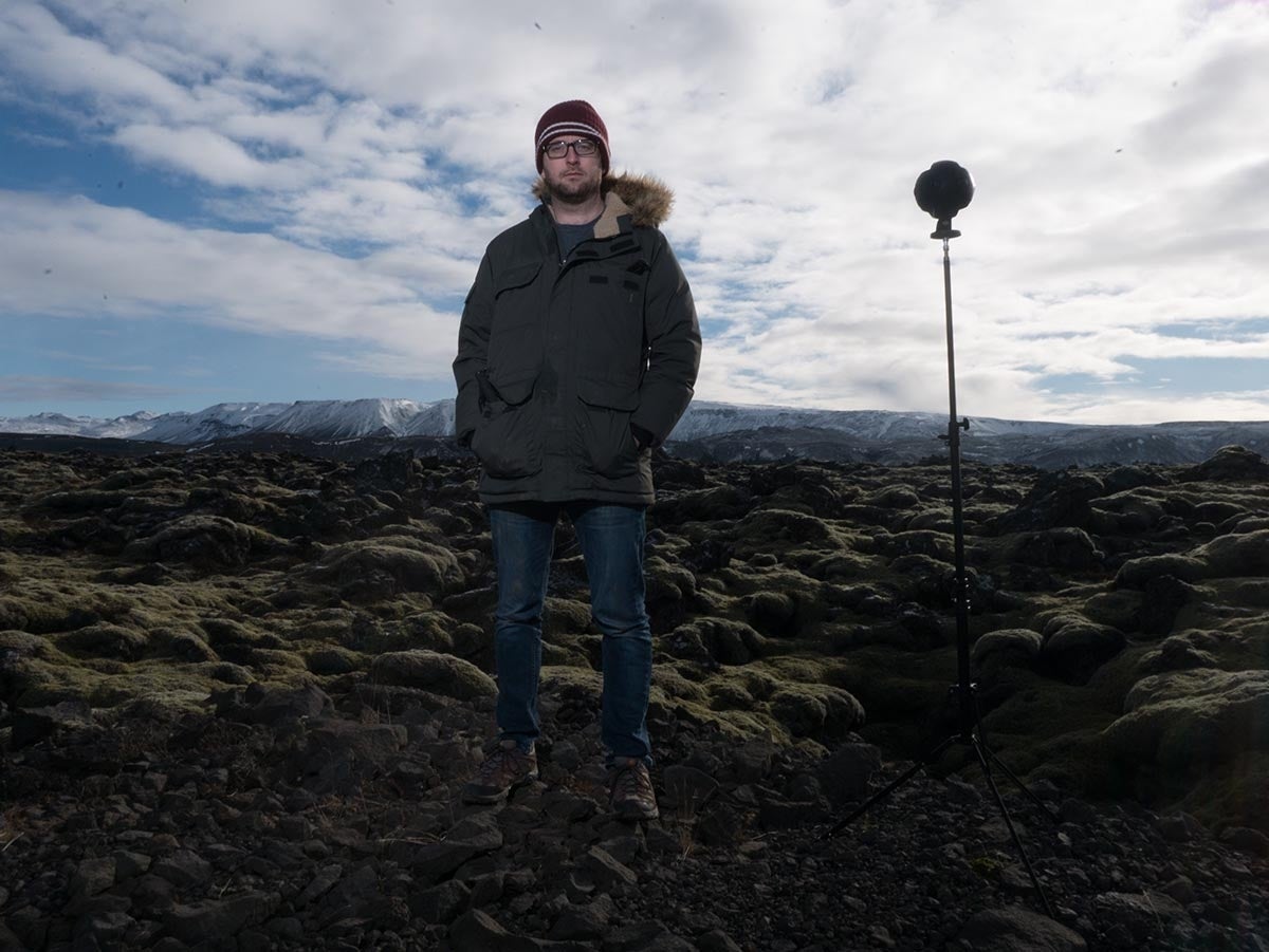 Man in a parka and hat standing in a wide open landscape with a VR camera on a stand