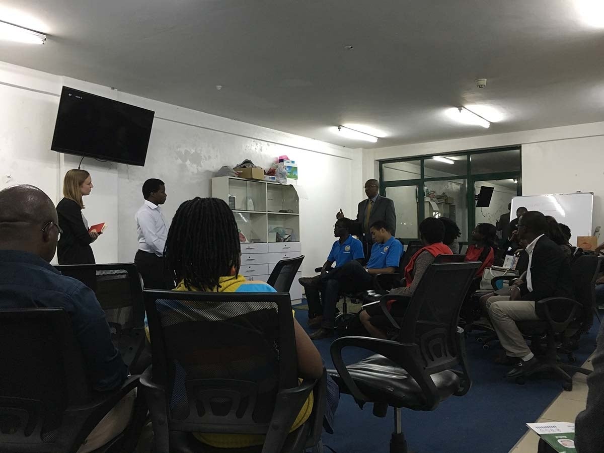 Group of people seated paying attention to three standing people at the front of the room who are speaking.