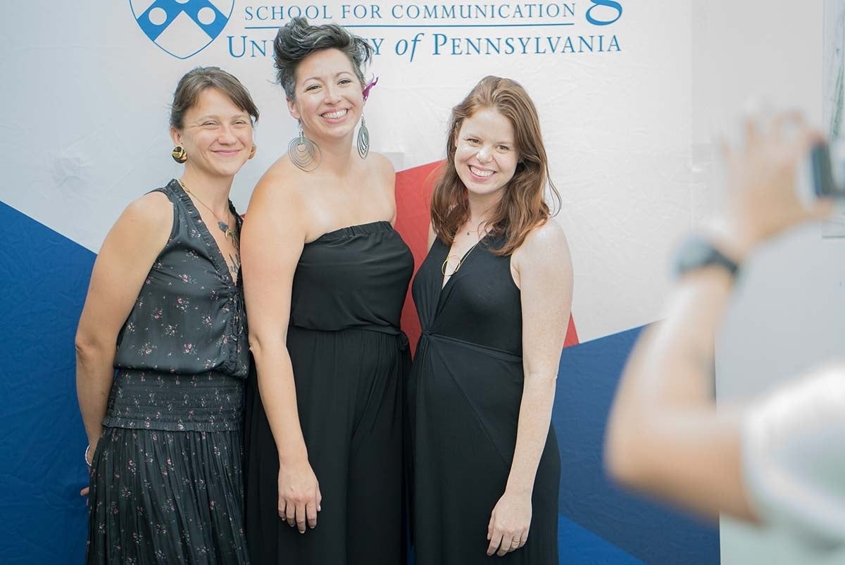 Emily LaDue, Lyndsey Beutin, and Corrina Laughlin posed for a picture in front of a white backdrop that has the Annenberg School for Communication's logo and red and blue shapes.