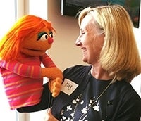 Amy Jordan facing and smiling at a muppet. Muppet has orange hair, blue eye shadow, eyeliner, a red nose, and a pink shirt with blue and yellow stripes