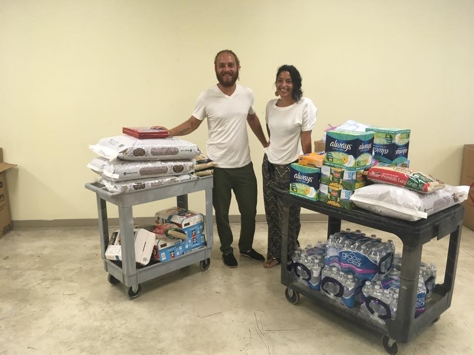 Luis Rodríguez and Christine Nieves Rodríguez posing with supplies on gray and black rolling carts. Supplies such as sanitary napkins, rice, Quaker Oats products and packs of water are on the carts. 