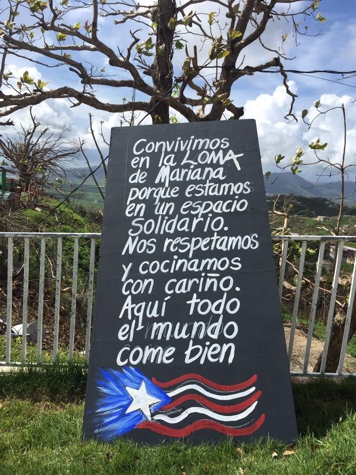 Chalkboard that says in Spanish "Convivimos en la LOMA de Mariana porque estamos en un espacio solidario. Nos respetamos y cocinamos con cariño. Aquí todo el mundo come bien' and below message is Puerto Rican flag drawn with chalk.