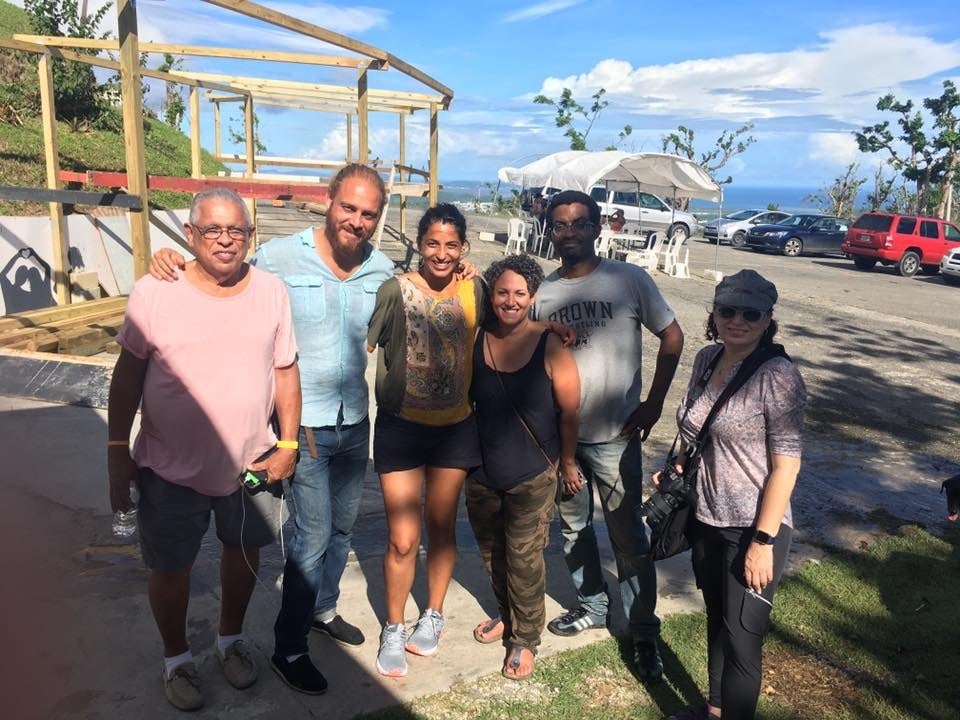 Rodríguez Sanchez and Nieves Rodríguez with Carmen Maldonado and other volunteers. They pose in font of a wooden frame for some structure, and a white tent with tables and chairs under it. The sky is blue and there are clouds. They are by water. 