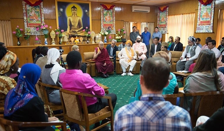 In a circle formation, attendees are seated and facing the Dalai Lama who sits at the top of the circle. The Dalai Lama is speaking to the group. 