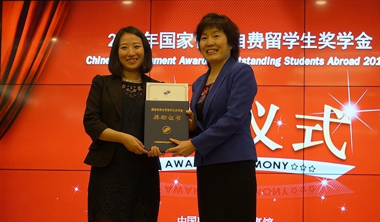 Jingwen Zhang receives her award from Ambassador Zhang Qiyue. They are in front of a digital screen that has a red background with a design for the awards show. 