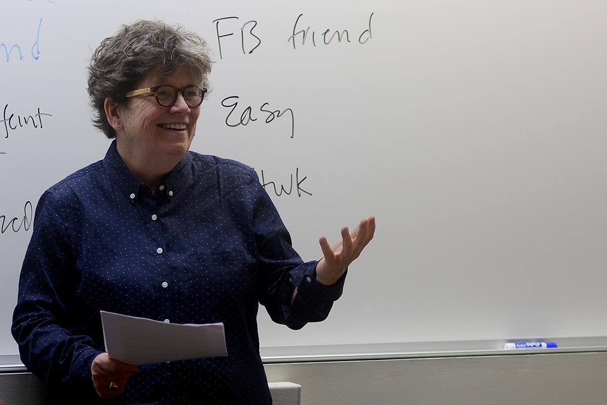 Jaoan Garry standing and speaking in front of a whiteboard with some words written it. She is holding papers in her right hand. 