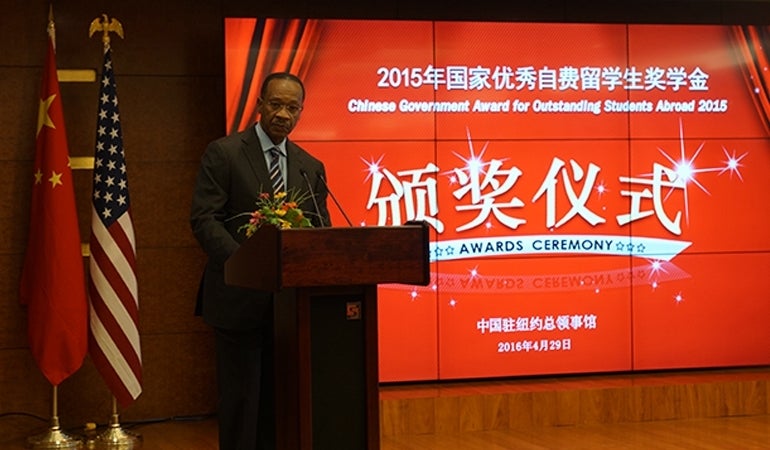 John Jemmott delivers a speech while standing behind a podium. Behind are are the Chinese and American flags beside each other. To his left is the digital screen for the awards show. 
