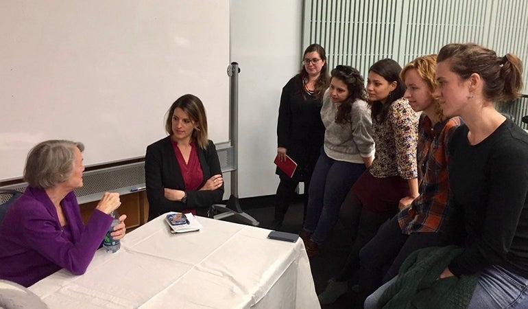 Ferrari and other MARC scholars speaking with Bonnie Raines. Raines is seated behind a table with a white cloth, and those listening are in front of her standing or leaning on a desk.