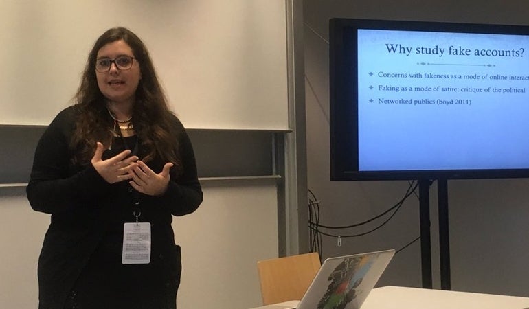 Elisabetta Ferrari standing while making a presentation. To her left, she has a presentation open. The slide is titled "Why study fake accounts?" and has three bullet points.