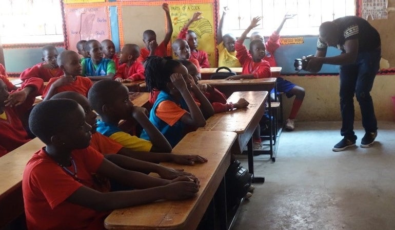 The setting is a classroom from a side-view, and there are many students seated facing towards the front and side. Five students at the side have their hands raised. At the front is a man videoing the children. 