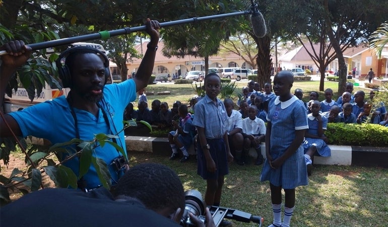 Two school children in Kampala interview one another on camera. A man in a blue shirt and headphones holds up the microphone in front of them and a camera man in a navy blue shirt is crouched beside him. Behind the two children are more children sitting down. They are all outside and there are trees in the background as well.