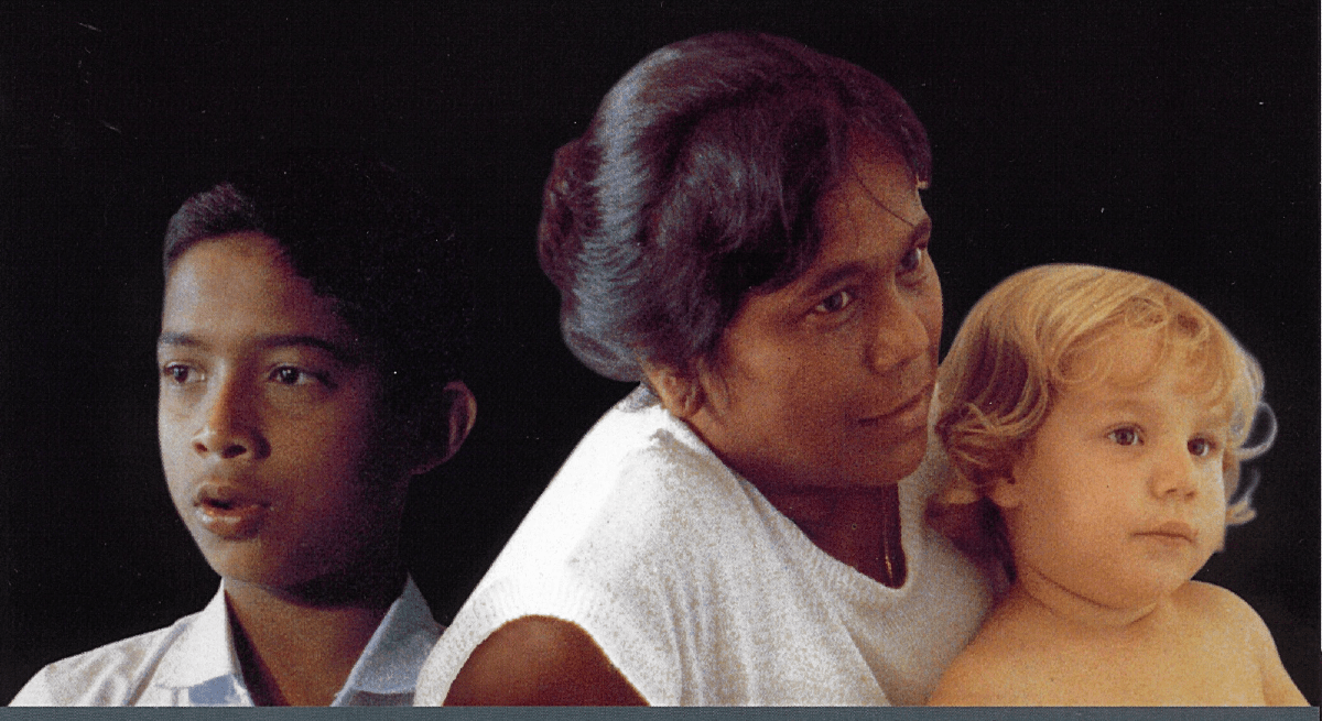 The background is all black. The foreground has 3 people. One is a young child who is on the left of the picture and is looking in that direction. The other two are an Indian woman and a white baby in her lap. They are to the right of the image and face that way.
