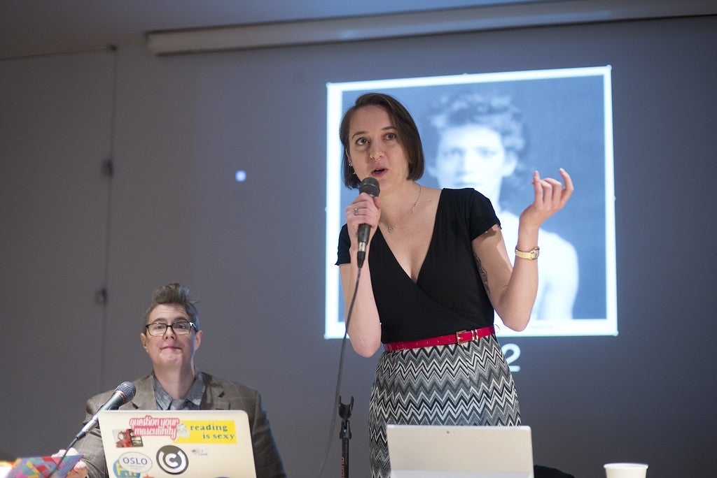 Jessa Lingel standing and speaking into a wired microphone and Jack Gieseking sitting beside her, making eye contact with the camera. They are both behind a table with laptops open. Behind them is a projected black and white image of someone. 