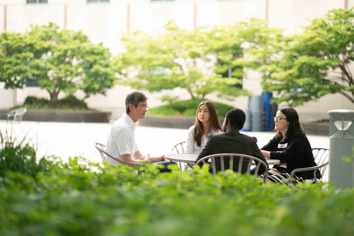 Four people sitting around a metal table in the mid-ground, with some outdoor shrubbery visible